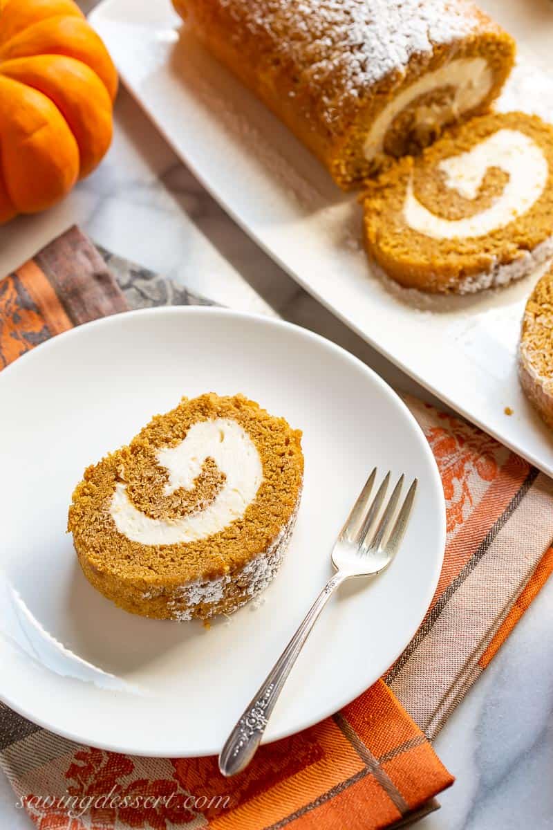 A plate with a slice of pumpkin roll next to a platter with the remaining pumpkin roll