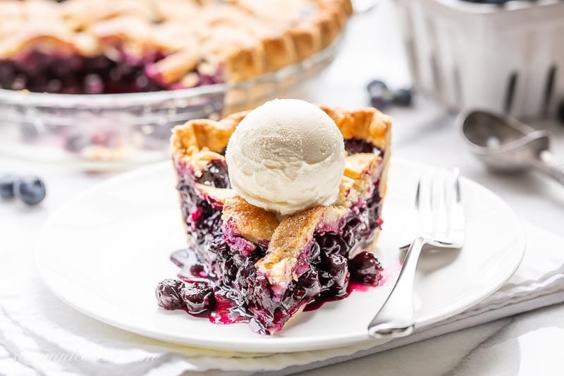 A slice of pie topped with vanilla ice cream and blueberries spilling out onto a plate