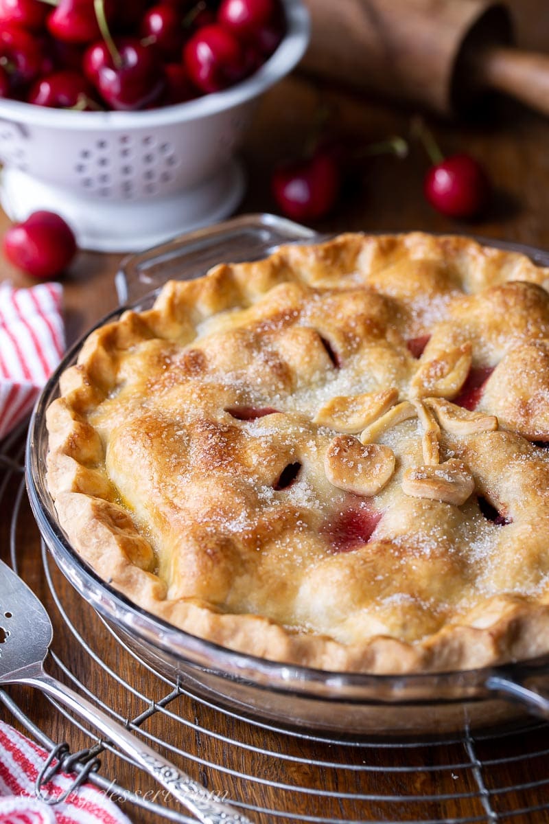 A homemade double crust pie on a cooling rack with fresh cherries in the background