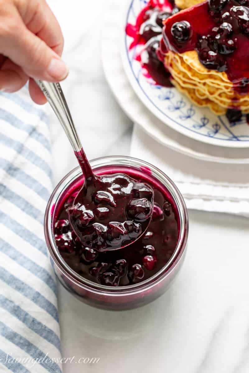 a jar of blueberry sauce with a spoonful being scooped out with pancakes in the background
