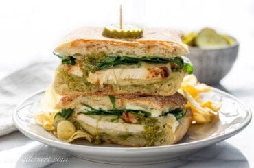 A stacked chicken pesto panini sandwich topped with a pickle slice.