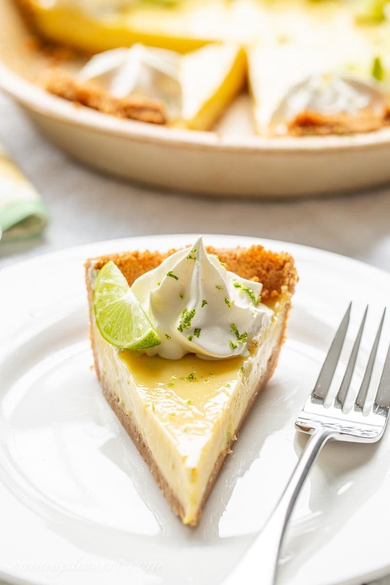 A slice of key lime pie on a plate garnished with key lime wedges, whipped cream and lime zest