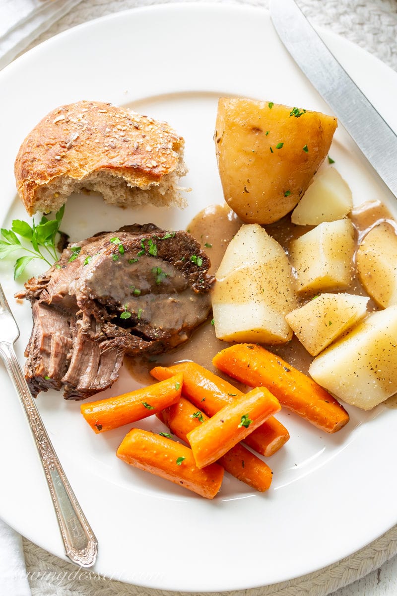 A dinner plate with slow cooker pot roast covered in gravy with roasted potatoes, carrots and crusty bread