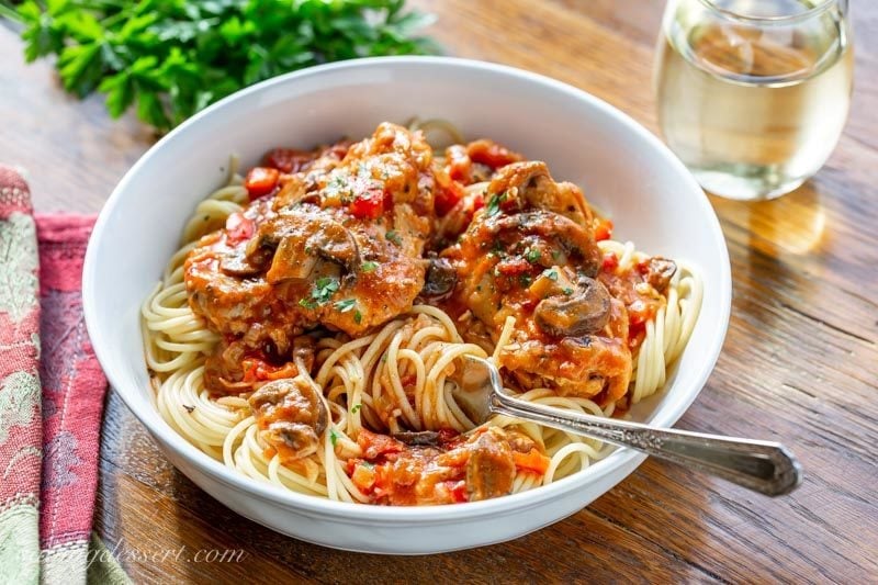 A bowl of spaghetti topped with Chicken Cacciatore with mushrooms, red peppers and parsley served with a glass of white wine