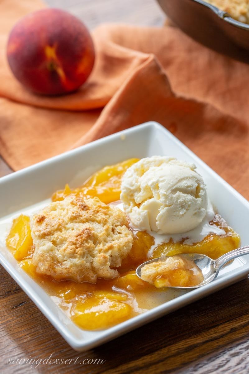 A bowl with old fashioned peach cobbler with a shortcake biscuit crust and a scoop of vanilla ice cream