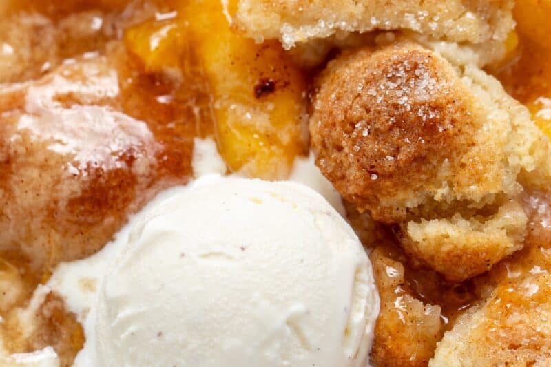 A closeup photo of a bowl of peach cobbler with a scoop of ice cream on top.