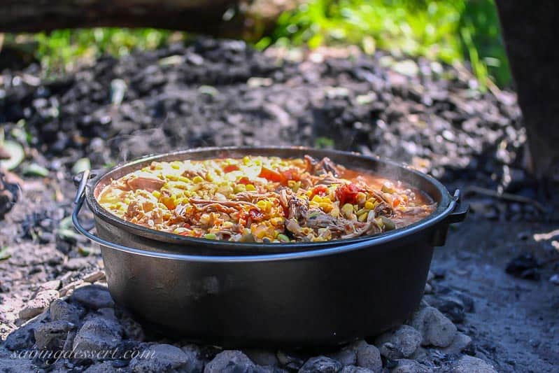 A cast iron Dutch oven filled with stew over an outdoor fire with charcoal.