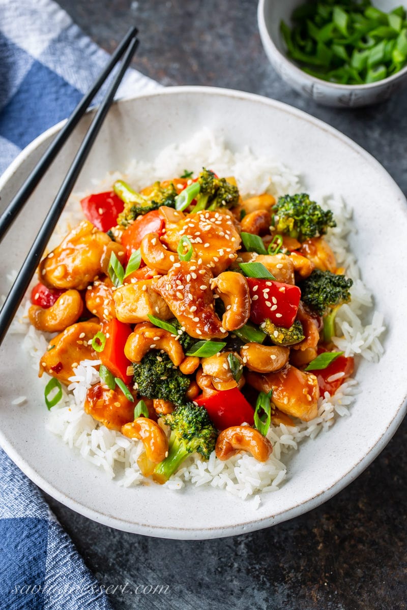 Honey Cashew Chicken - You'll love the sweet heat of this Asian inspired stir-fry loaded with tender chunks of chicken, red bell pepper, onions & tender broccoli. #savingroomfordessert #honeycashewchicken #cashewchicken #Asianstirfry #stirfry #chickenstirfry #chicken #Sriracha #cashew #dinner #weeknightmeal A bowl of Jasmine rice topped with Honey Cashew Chicken with red bell pepper, broccoli, onions and cashews. Garnished with sesame seeds and sliced green onions.