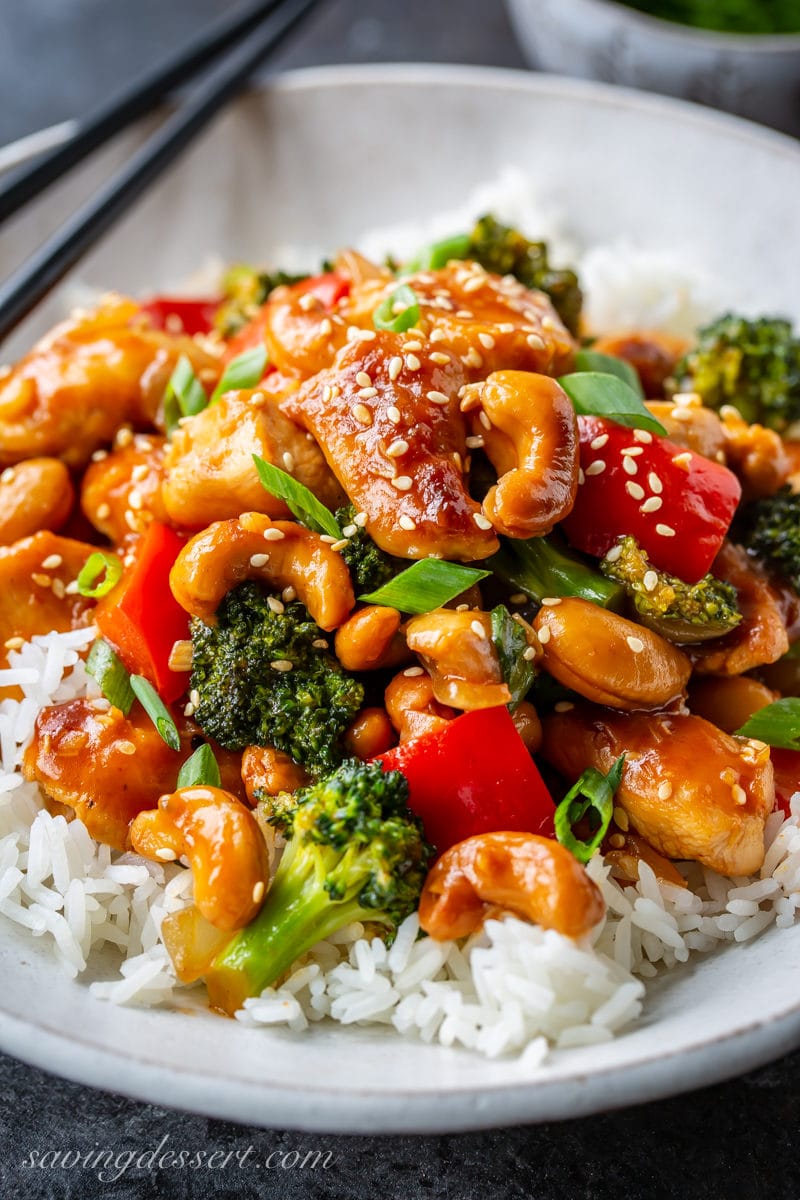 Up close view of a bowl of Jasmine rice topped with Honey Cashew Chicken with red bell pepper, broccoli, onions and cashews. Garnished with sliced green onions and sprinkled with sesame seeds.