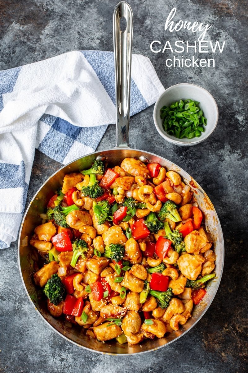 A skillet with Honey Cashew Chicken with red bell pepper, broccoli and onions garnished with sliced green onions and sesame seeds.