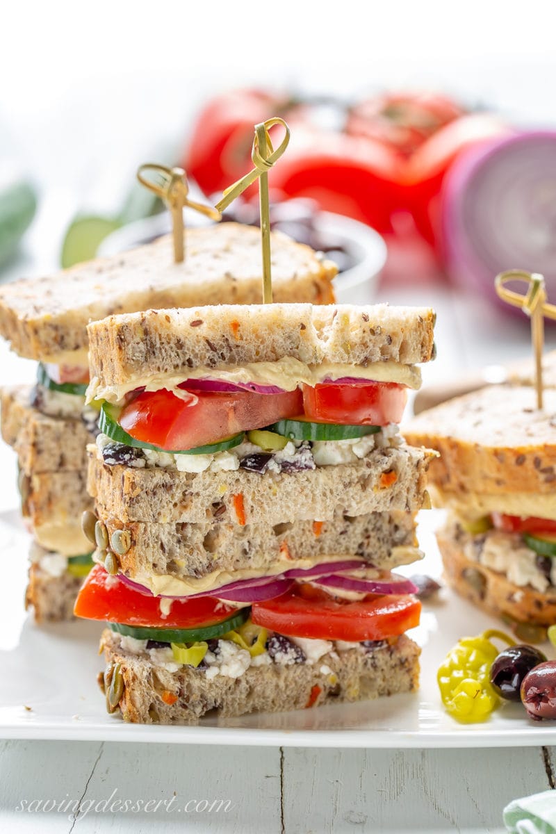 This might look like a plain old veggie sandwich, but it's so much more! The Greek flavors are bright, briny, crisp, fresh and delicious! #hummussandwich #greeksaladsandwich #greeksandwich #vegetariansandwich #makeaheadsandwich #picnicsandwich #greekfood A stack of Greek Salad Hummus Sandwiches with cucumbers, tomatoes, onion, feta and olives on a nice seeded bread.