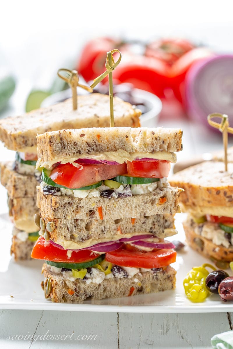 A stack of Greek Salad Hummus Sandwiches with cucumbers, tomatoes, onion, feta and olives on a nice seeded bread.