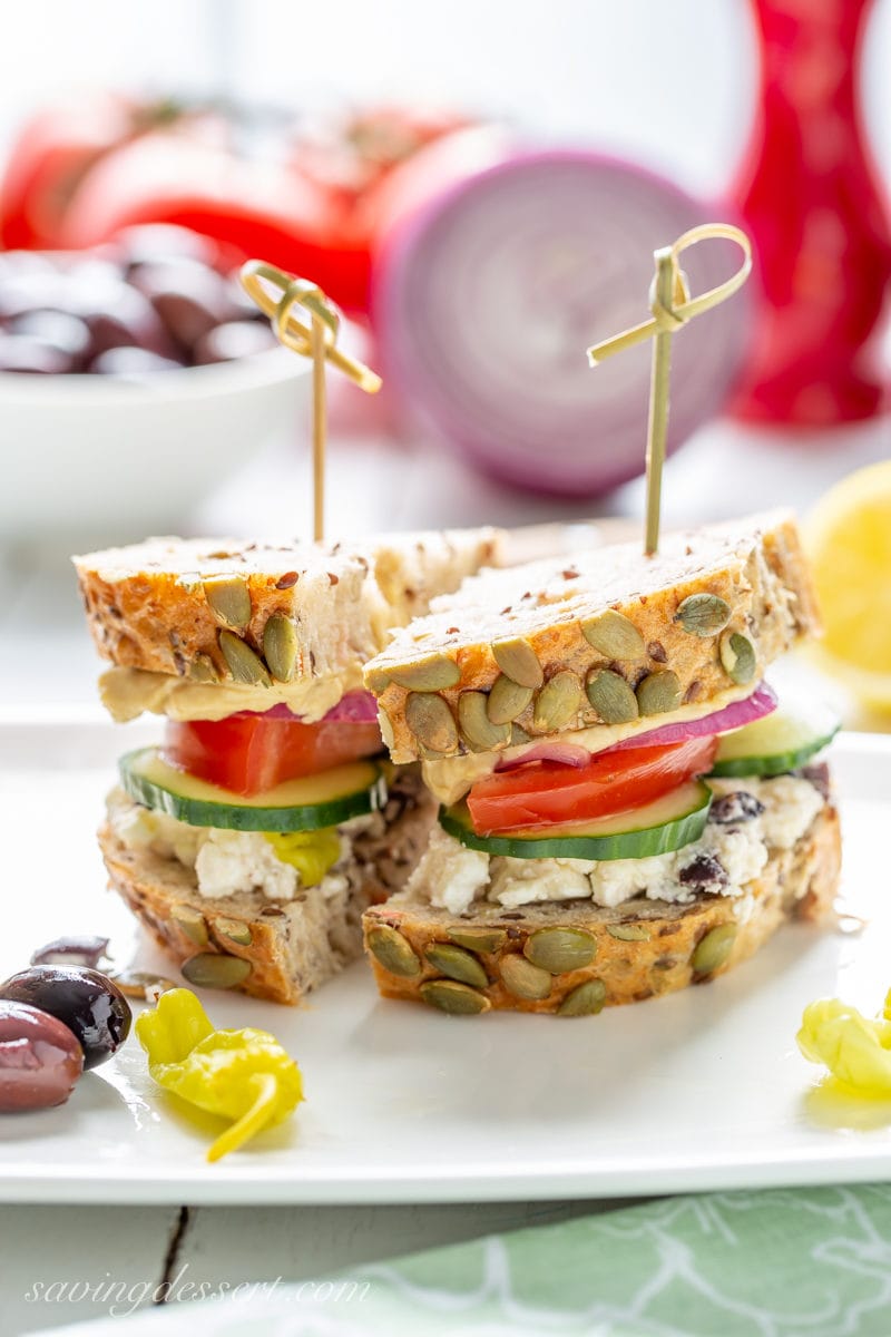 A Greek Salad Hummus Sandwich sliced in half served with peppers and olives