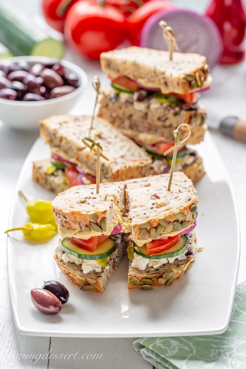 A tray of sliced Greek Salad Hummus Sandwiches served with olives and peppers.