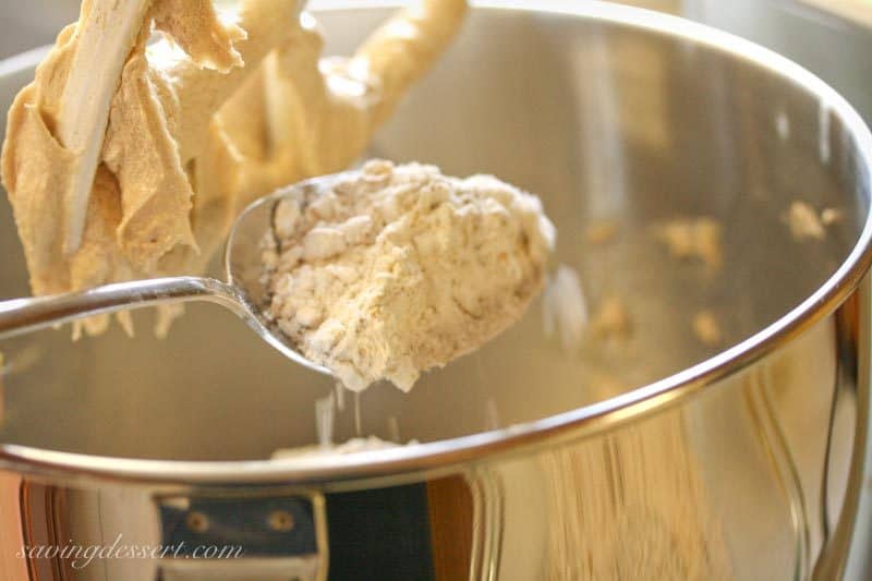 A mixing bowl filled with cookie dough with a spoonful of flour being added