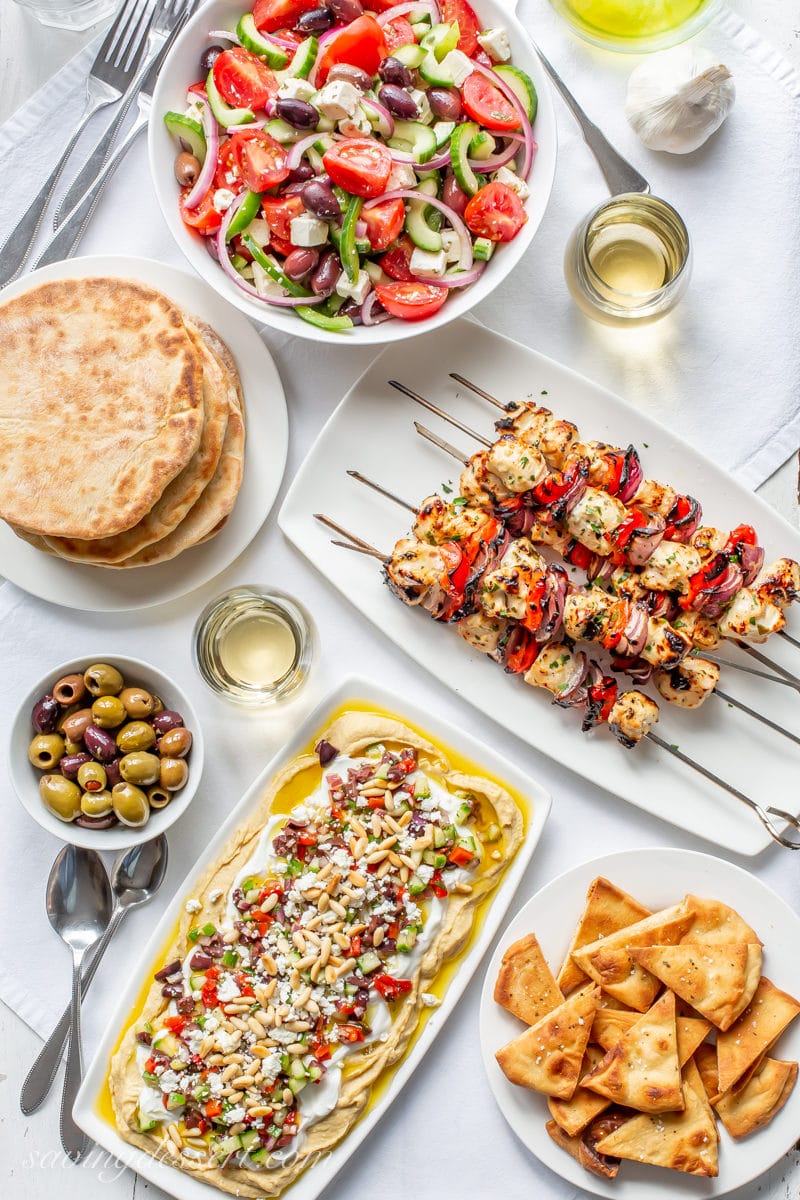 A table filled with Greek inspired recipes from homemade whole wheat pitas, Greek Salad and hummus dip with olives