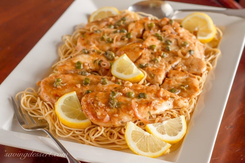 Try this classic Chicken Piccata Recipe made with tender chicken cutlets quickly sautéed then simmered in a light lemony, white wine sauce. Serve over pasta with capers and lemon wedges for an easy weeknight meal. #savingroomfordessert #chicken #chickenpiccata #easypiccata #chickenpiccatarecipe #chickencutlets #weeknightdinner #italian A platter of chicken piccata with lemon wedges, capers and shallots. Served over spaghetti