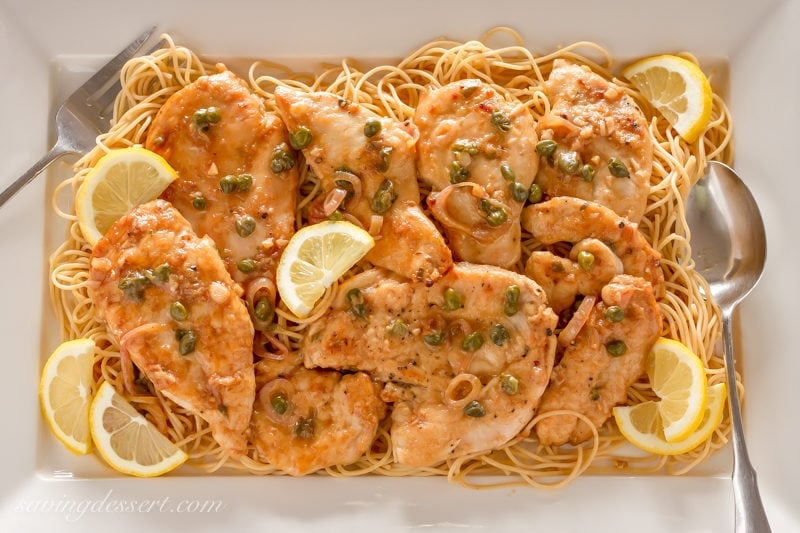 Try this classic Chicken Piccata Recipe made with tender chicken cutlets quickly sautéed then simmered in a light lemony, white wine sauce. Serve over pasta with capers and lemon wedges for an easy weeknight meal. #savingroomfordessert #chicken #chickenpiccata #easypiccata #chickenpiccatarecipe #chickencutlets #weeknightdinner #italian A platter of Chicken Piccata over spaghetti garnished with lemon wedges