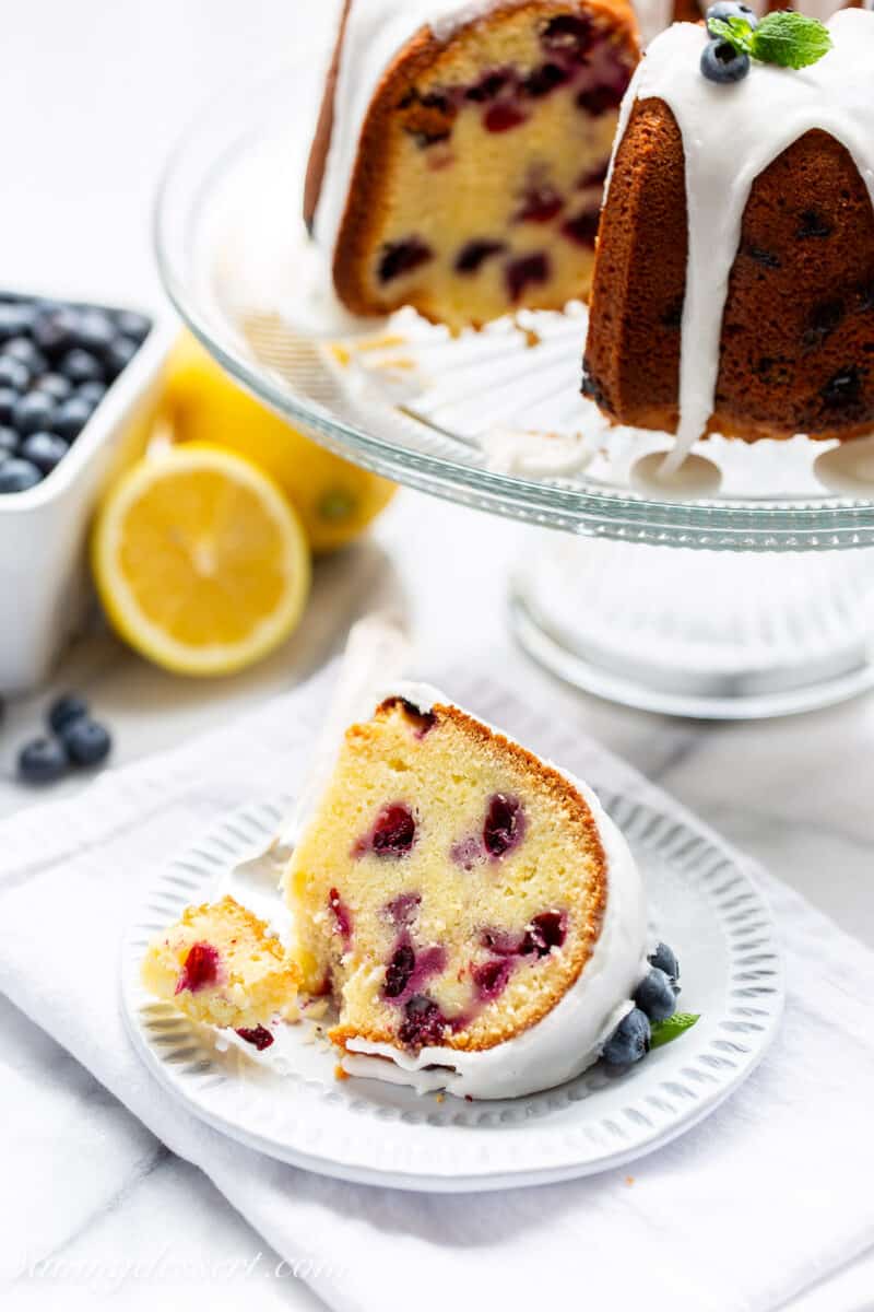 A slice of blueberry pound cake on a plate with a fork and blueberries and lemon in the background.