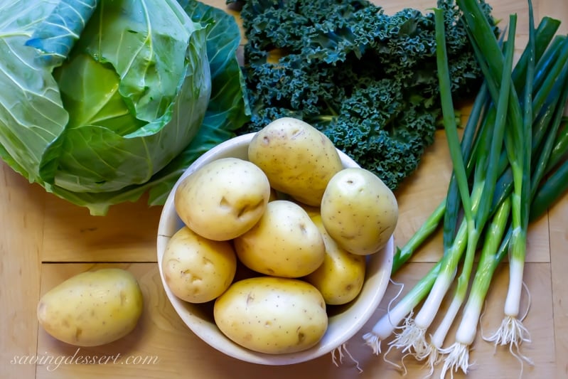 A cutting board with potatoes, cabbage, kale and scallions