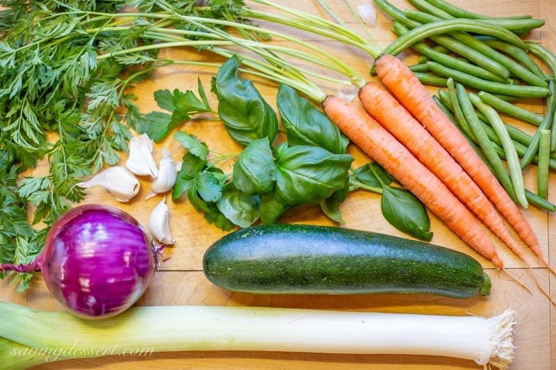 fresh carrots, green beans, a zucchini, garlic cloves, a red onion and a large leek piled on a cutting board with parsley and basil