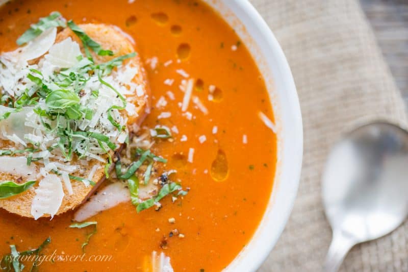 A bowl of fresh roasted tomato soup with a slice of toast on top with Parmesan and basil leaves