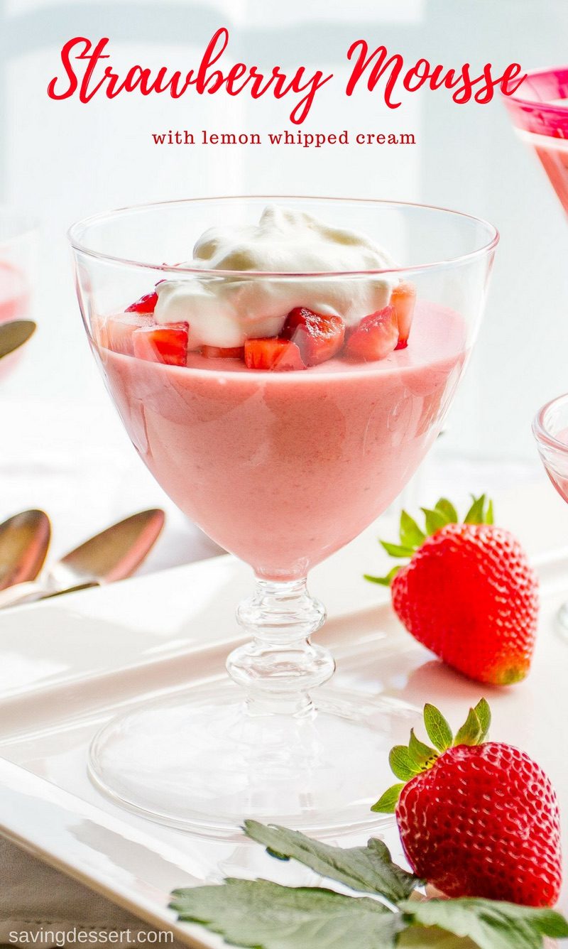 A clear glass stemware glass filled with bright pink strawberry mousse topped with diced strawberries and whipped cream.