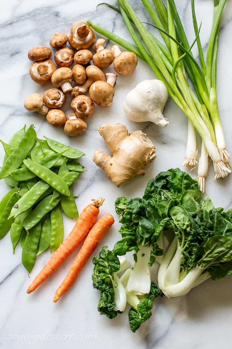 Various vegetables on a marble countertop including carrots, mushrooms, snow peas, green onions, garlic and bok choy