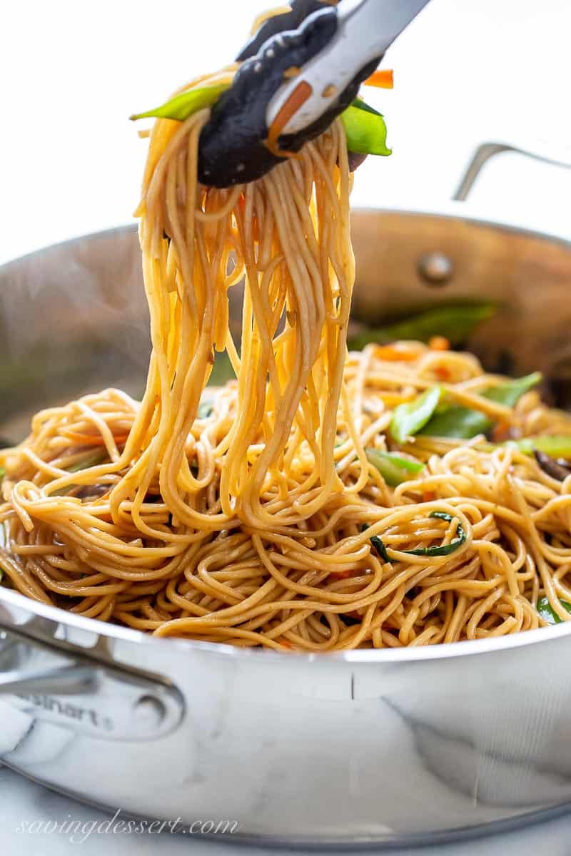 Tongs picking up lo mein noodles from a skillet