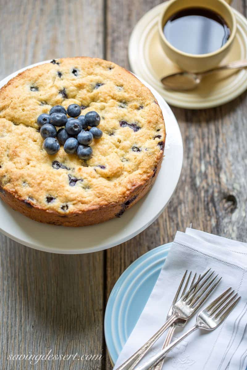 A blueberry breakfast cake on a cake stand and a cup of coffee