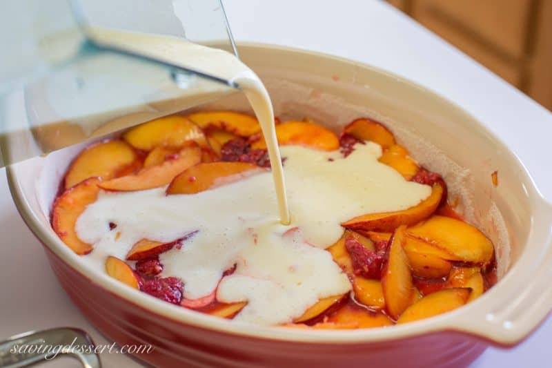 A casserole filled with peaches and raspberries with a custard mixture being poured over the fruit.