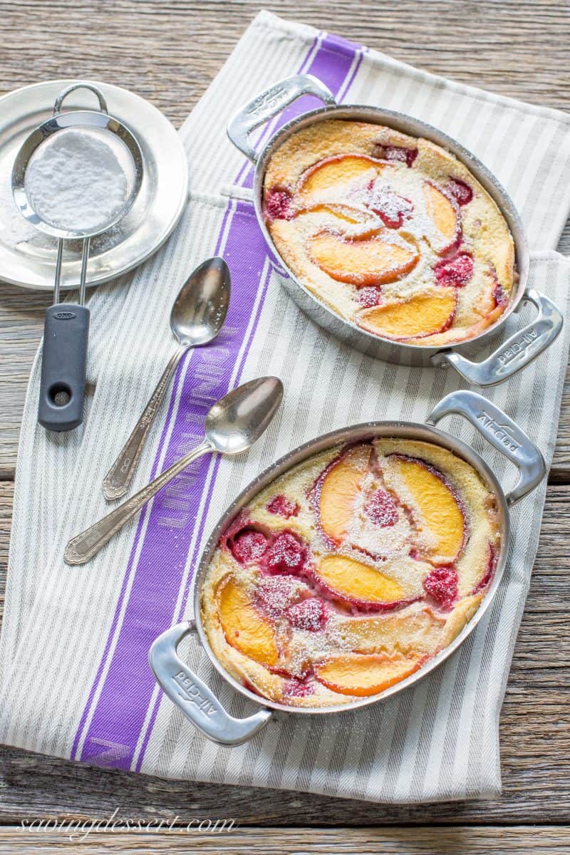 An overhead photo of two small casserole dishes filled with fresh peach and raspberry clafoutis dusted with powdered sugar.