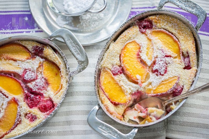 An overhead shot of two small casserole dishes with a peach and raspberry clafoutis dusted with powdered sugar.