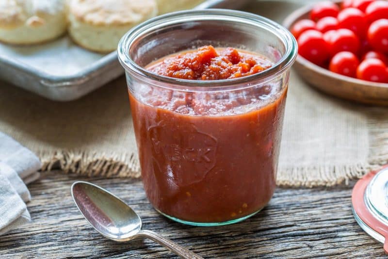 A jar of tomato jam with biscuits in the background.
