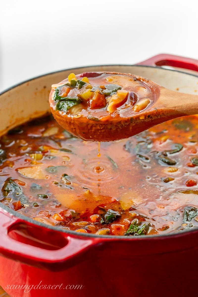 A Dutch oven filled with vegetable soup with a ladle hovering over the pot dripping broth