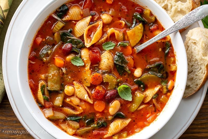 A close up of a bowl of vegetable pasta soup with spinach and fresh basil leaves