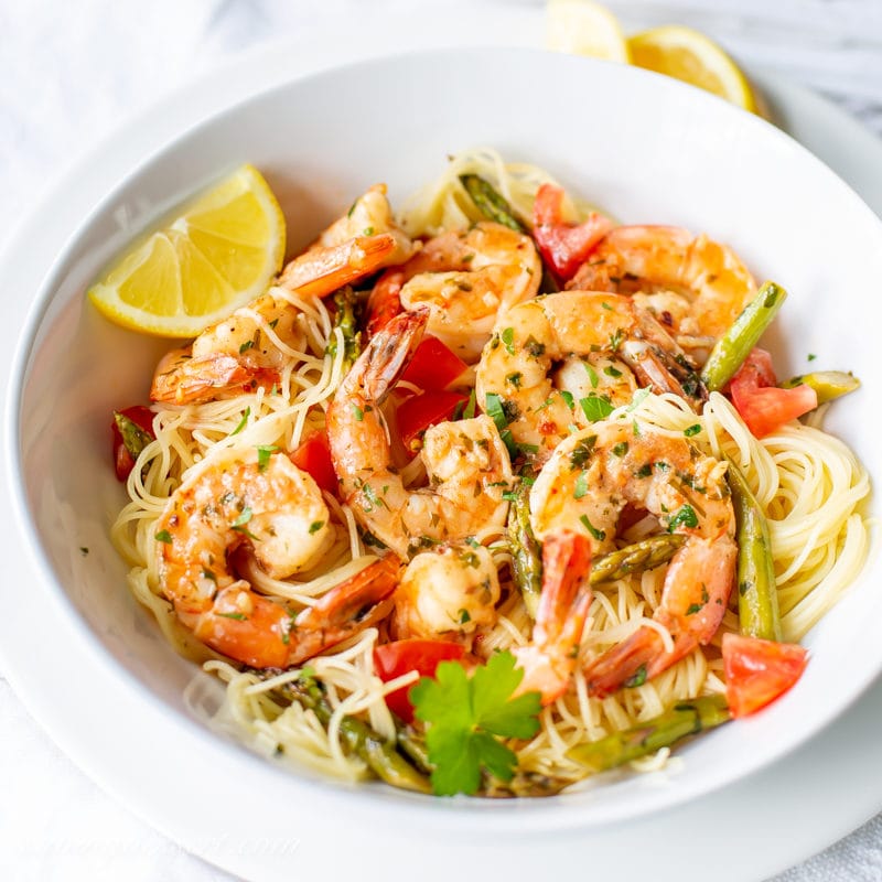 A bowl of shrimp scampi with tomatoes, asparagus and lemon wedges