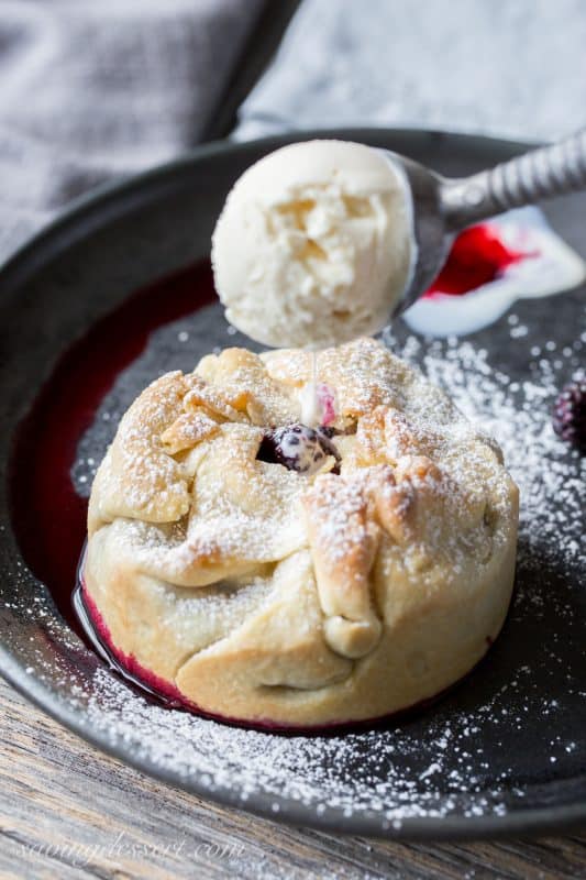 A deep dish mini black raspberry pie on a plate being topped with a scoop of vanilla ice cream