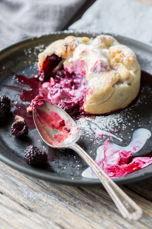 A mini berry pie opened with a spoon with berries and juices pouring onto the plate
