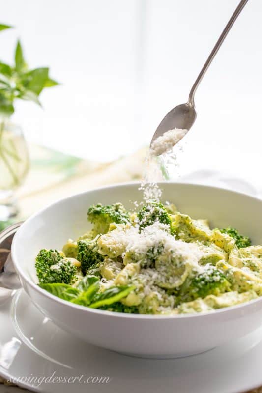 A bowl of pasta in basil cream sauce with fresh Parmesan being tossed over the top