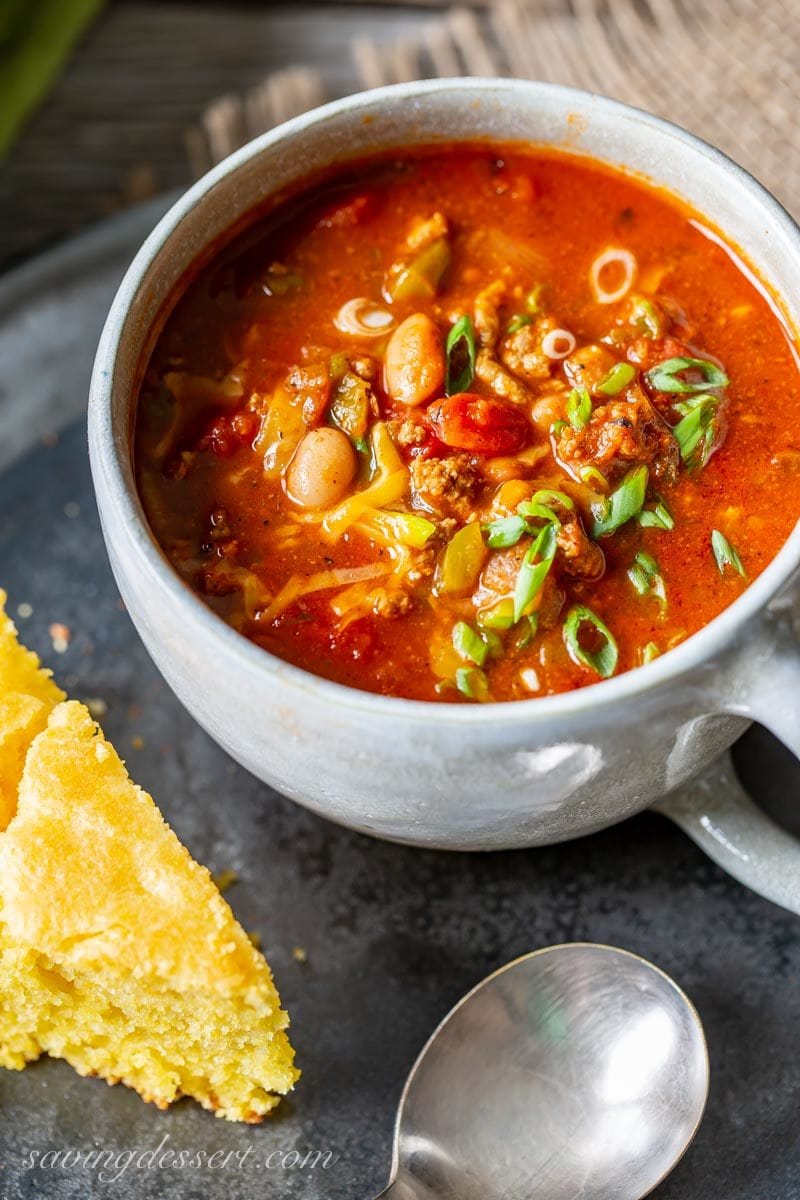 A mug of chili topped with cheddar cheese and sliced green onions served with cornbread