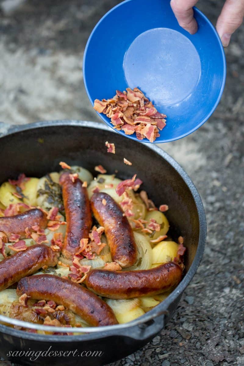 A camping Dutch oven filled with potatoes, onions and bangers topped with bacon