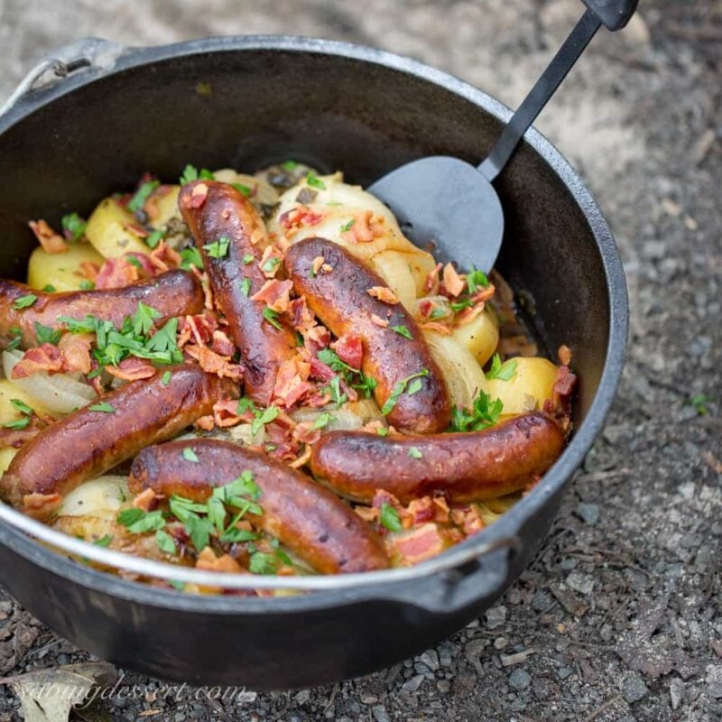 An outdoor Dutch oven filled with potatoes, onions and sausages topped with bacon and parsley