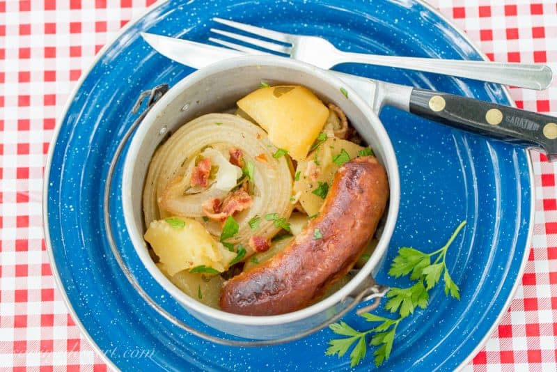 A plate with a camping pot filled with bangers onions and potatoes with bacon on top