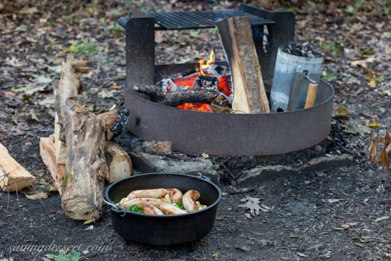 A campfire ring with hot coals and fire wood and a Dutch oven filled with sausages and potatoes
