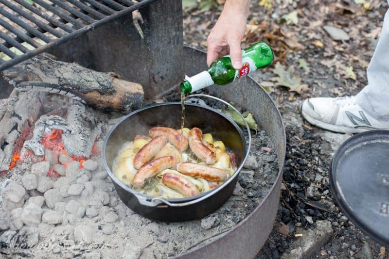 A camping Dutch oven filled with sausages, potatoes and onions with a bottle of beer being poured inside