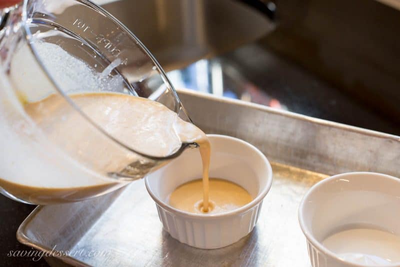 A measuring cup pouring custard into small ramekins set inside a baking pan