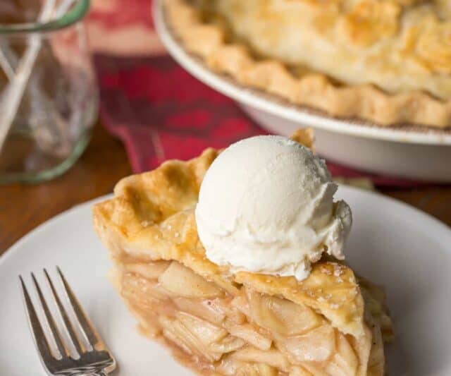 Front view of a slice of apple pie on a plate topped with a scoop of ice cream.