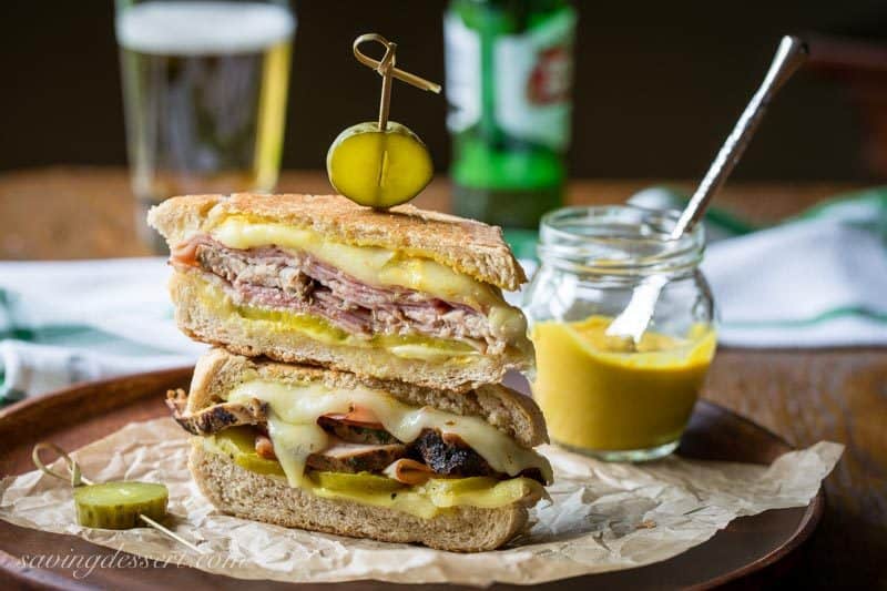 A side view of a stacked cuban sandwich topped with pickles and served with beer