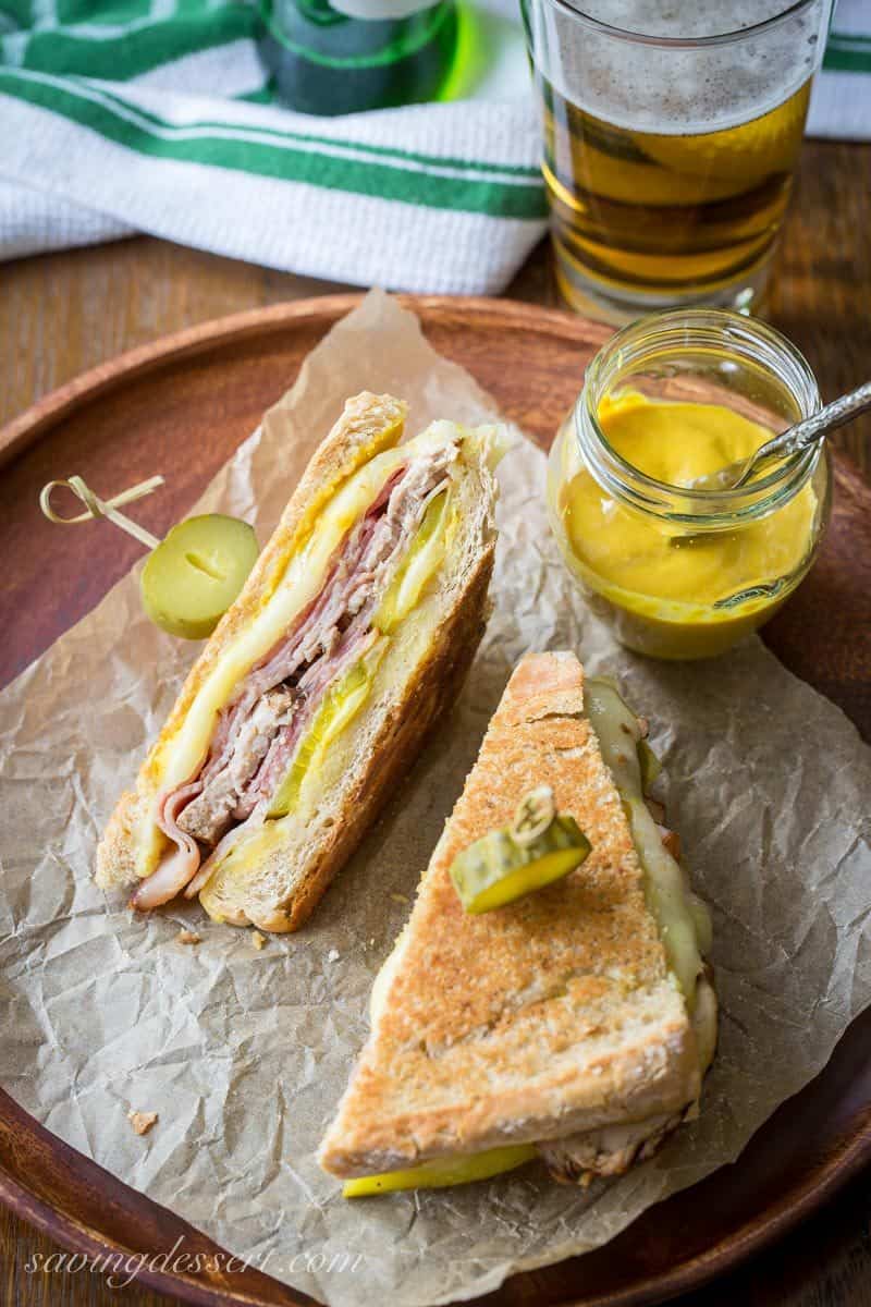 An overhead view of a toasted ham and pork sandwich with mustard and beer on the side